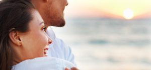 A young couple, a man and a woman, laughing together while standing on a sunny beach, enjoying a joyful moment by the ocean.